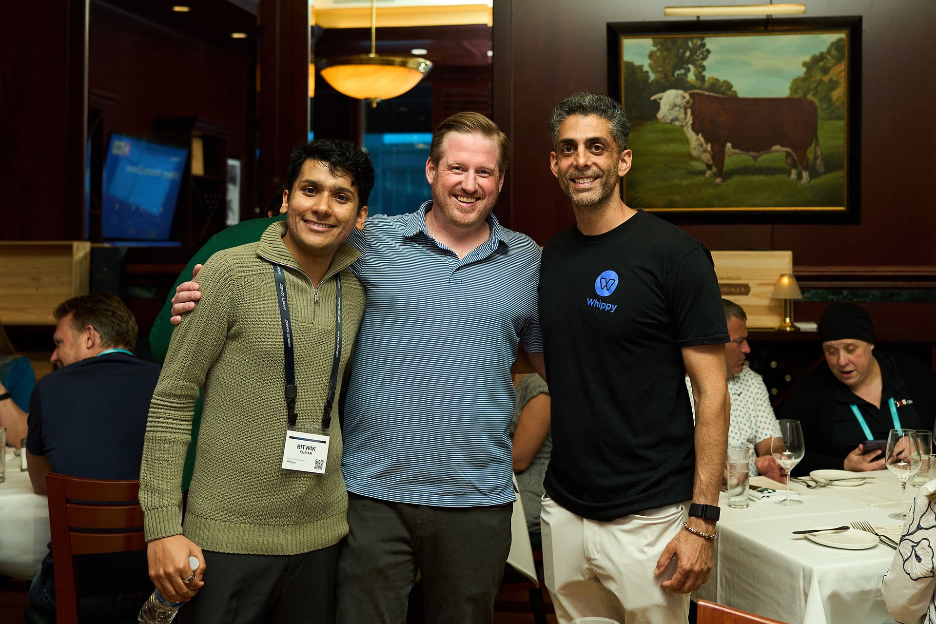 Three men smiling together at the Avionté CONNECT 2026 dinner, one wearing a Whippy branded t-shirt and another with an Avionté CONNECT lanyard